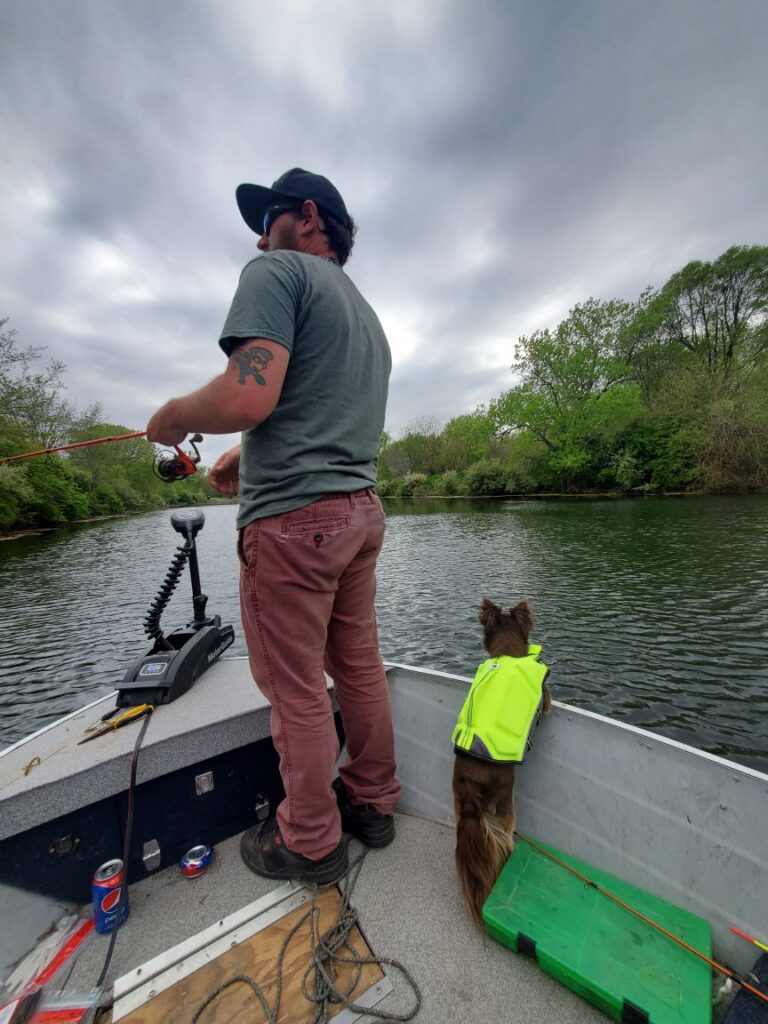 person standing in boat fishing while pet cat wearing safty vest looking overboard at the water